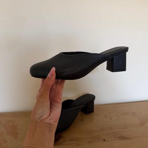May include: A pair of black mules featuring a smooth, matte finish, a rounded toe, and a sturdy block heel. The slip-on design has a clean, unadorned upper that covers the front of the foot, providing a simple and functional footwear option.