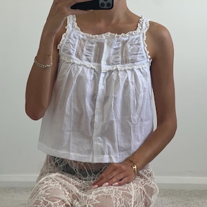 May include: A white sleeveless top featuring a square neckline, ruffled shoulder straps, and a button-down front. The bodice includes vertical panels of intricate lace detailing and gathered fabric, creating a textured, lightweight appearance.