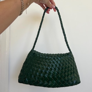 May include: A dark green woven handbag with a plaited handle. The bag has a rectangular shape with a slightly curved top. The woven pattern creates a textured surface. The bag is held up by a hand.