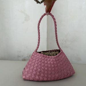 Può includere: Una borsa a mano rosa caratterizzata da un motivo esterno intrecciato simile a un cesto e da un unico manico intrecciato. La borsa ha una forma trapezoidale strutturata e include una chiusura a coulisse in tessuto beige visibile all'apertura.