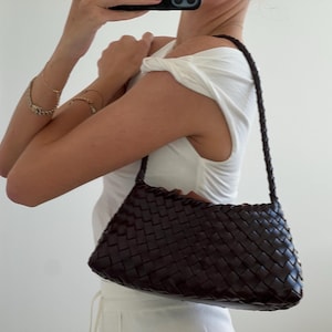 May include: A dark brown, woven leather shoulder bag with a braided strap. The bag has a rectangular shape and a textured surface. The bag is held by a person wearing a white top.