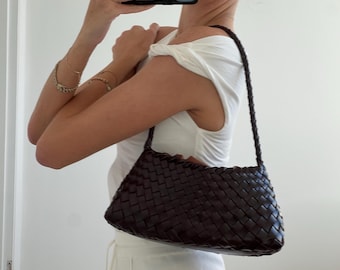 May include: A dark brown, woven leather shoulder bag with a braided strap. The bag has a rectangular shape and a textured surface. The bag is held by a person wearing a white top.