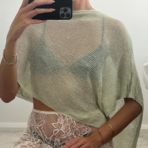 May include: A light green, sheer, knitted poncho-style top. The top is worn over a black bra and paired with white lace pants. Gold bracelets adorn the wrist. The overall aesthetic is casual and stylish.