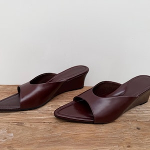 May include: A pair of dark burgundy leather slide sandals with a pointed toe and a low wedge heel. The sandals feature a wide strap across the top of the foot and a smooth, solid-coloured finish, providing a clean and simple design for footwear.