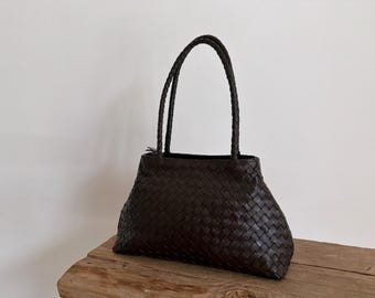 May include: A dark brown woven leather tote bag with braided handles. The bag has a trapezoidal shape and a spacious interior. The bag is sitting on a wooden surface.