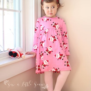 May include: A young girl wearing a pink dress with a Santa Claus print and pink tights. She is standing on a wooden crate and looking to the left. The dress has long sleeves and a gathered waist. The girl is wearing a red headband.
