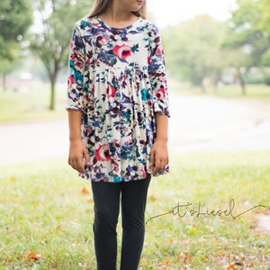 May include: A young person wearing a white dress with a floral print and black leggings. The dress has a gathered waist and 3/4 sleeves. The person is wearing brown boots.