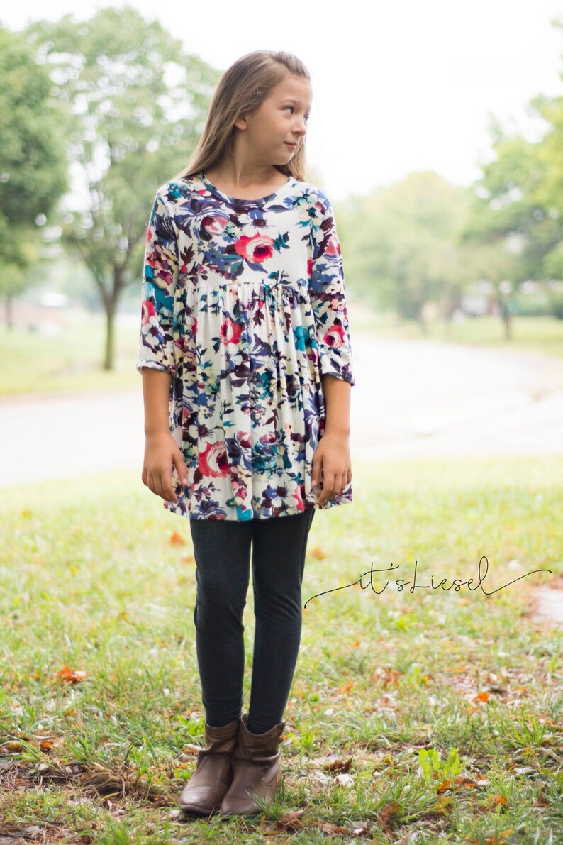May include: A young person wearing a white dress with a floral print and black leggings. The dress has a gathered waist and 3/4 sleeves. The person is wearing brown boots.