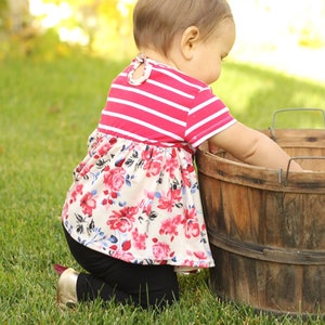 May include: A young child wearing a pink and white striped shirt with a floral print skirt and black leggings is kneeling in the grass next to a wooden barrel.