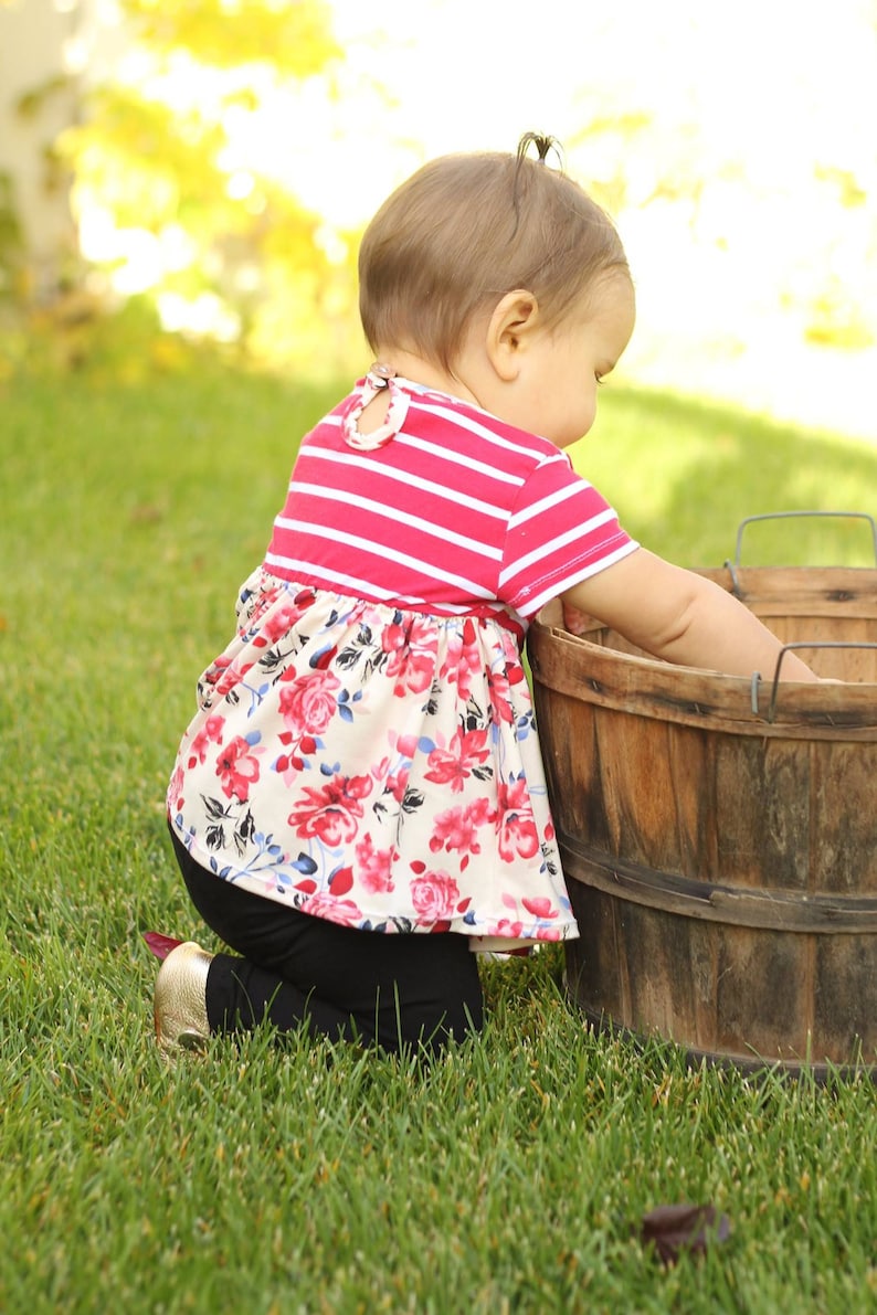 May include: A young child wearing a pink and white striped shirt with a floral print skirt and black leggings is kneeling in the grass next to a wooden barrel.