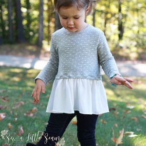 May include: A young girl wearing a grey and white polka dot top with a white ruffle bottom, black leggings, and black boots. She is standing in a grassy area with trees in the background.