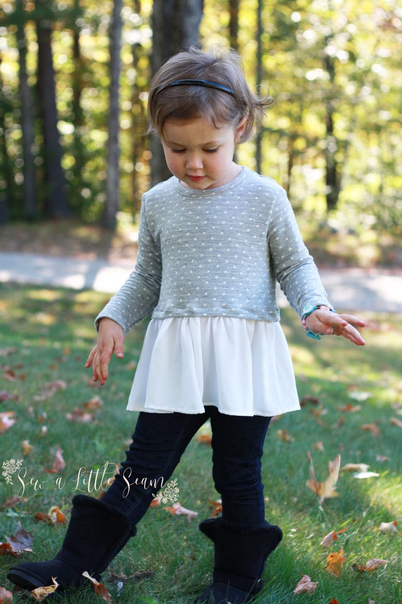 May include: A young girl wearing a grey and white polka dot top with a white ruffle bottom, black leggings, and black boots. She is standing in a grassy area with trees in the background.