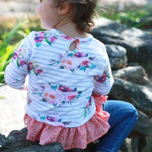 May include: A young child wearing a white and gray striped long-sleeved shirt with a floral print and a pink ruffled skirt. The child is sitting on a rock with their back to the camera.