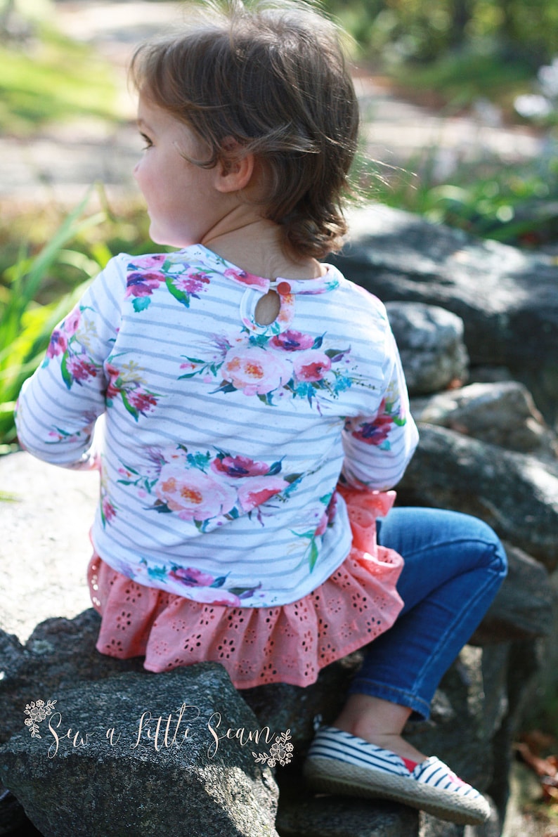 May include: A young child wearing a white and gray striped long-sleeved shirt with a floral print and a pink ruffled skirt. The child is sitting on a rock with their back to the camera.