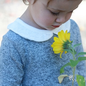 May include: A young child wearing a grey sweater with a white peter pan collar is smelling a yellow sunflower.
