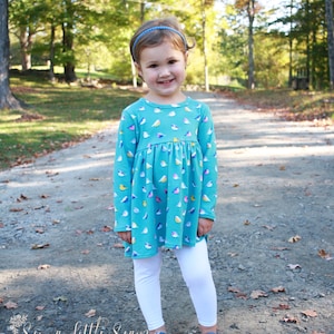 May include: A young girl wearing a teal blue long-sleeved dress with a colorful bird print. She is wearing white leggings and blue shoes. The girl is standing on a path in a wooded area.