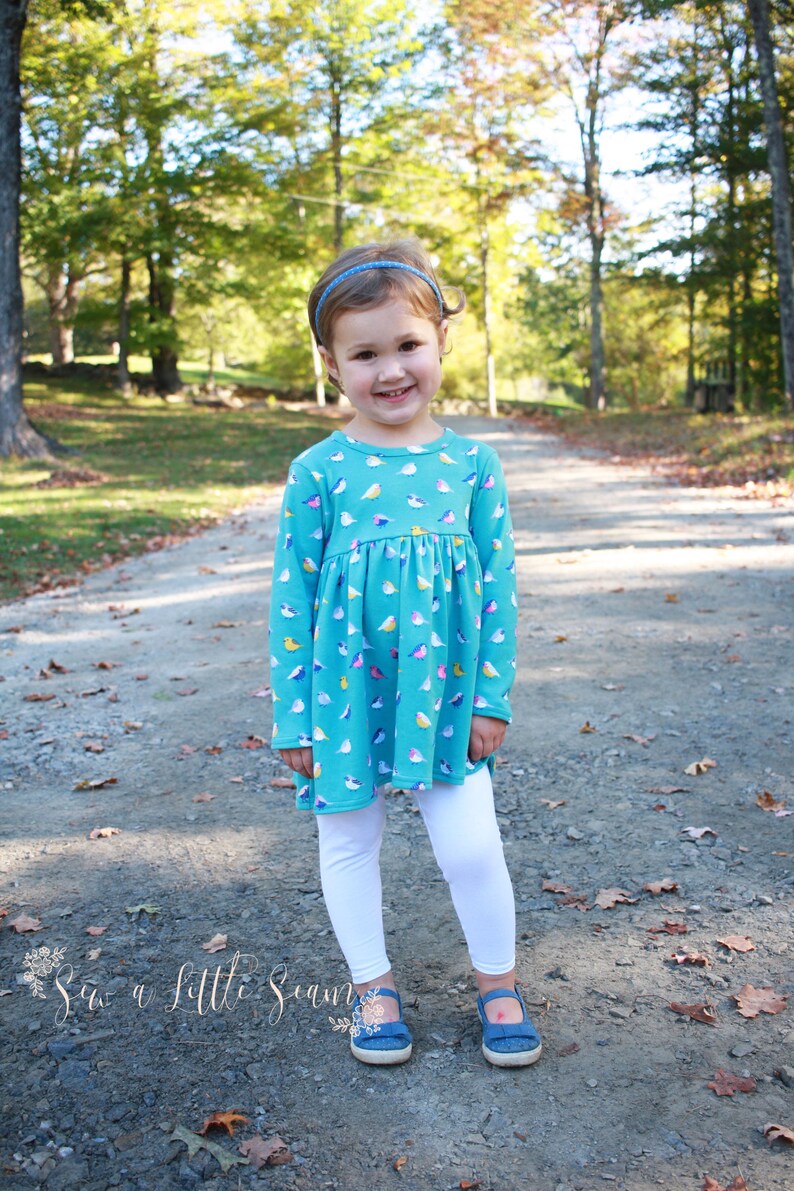 May include: A young girl wearing a teal blue long-sleeved dress with a colorful bird print. She is wearing white leggings and blue shoes. The girl is standing on a path in a wooded area.