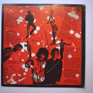 May include: Back cover of a vinyl record with a red and white splatter paint design. The record is titled "The Best of Jefferson Airplane" and features a photo of the band members. The track list is printed on the left side of the cover.