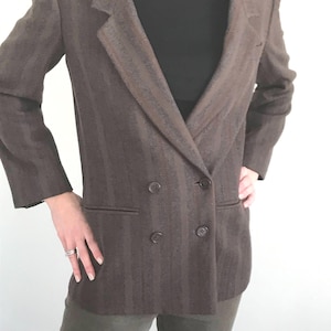 May include: A brown double-breasted blazer with vertical stripes. The blazer has two rows of buttons and two front pockets. The person is wearing a black turtleneck and olive green pants.