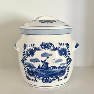 May include: A white ceramic jar with a lid, featuring blue hand-painted illustrations of a windmill in a landscape. The jar has two side handles and a decorative floral border surrounding the central scene, with blue accents on the lid and rim.