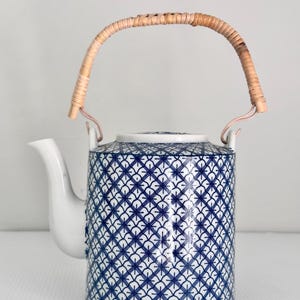 May include: A cylindrical ceramic teapot featuring a white base with a repeating blue geometric cross-hatch pattern. The teapot includes a curved white spout, a flat lid, and a flexible, light brown woven rattan handle attached to two loops.