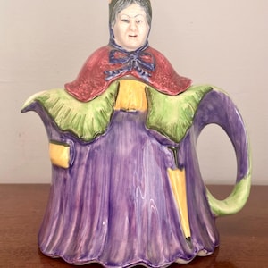 May include: A ceramic teapot shaped like a person wearing a dark bonnet, a red cape with a blue bow, a green shawl, and a long purple dress. The figure holds a yellow umbrella in one hand and has a yellow accent on the other side of the skirt.