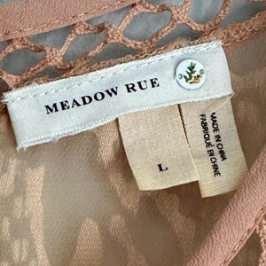 Może przedstawiać: Zbliżenie brzoskwiniowej metki odzieżowej z nazwą marki "MEADOW RUE" w czarnym tekście. Etykieta zawiera r&oacute;wnież rozmiar "L" oraz mniejszą metkę z napisem "MADE IN CHINA FABRIQUE EN CHINE". Odzież ma detal z szydełka.