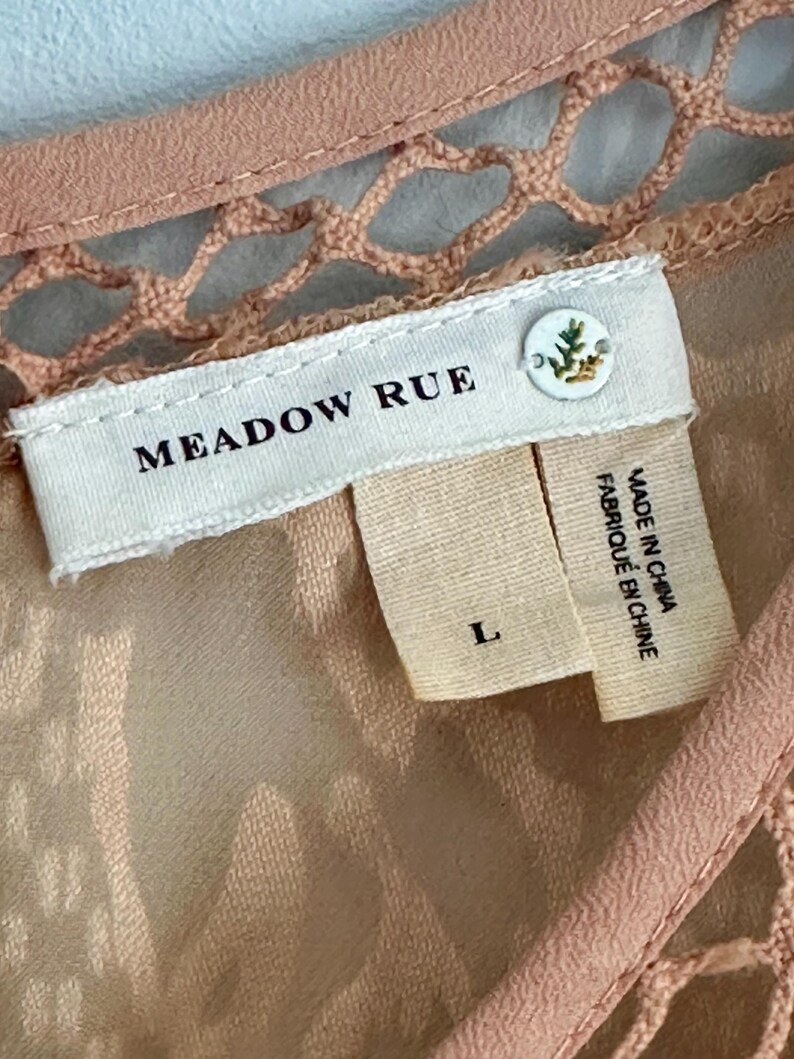 Może przedstawiać: Zbliżenie brzoskwiniowej metki odzieżowej z nazwą marki "MEADOW RUE" w czarnym tekście. Etykieta zawiera r&oacute;wnież rozmiar "L" oraz mniejszą metkę z napisem "MADE IN CHINA FABRIQUE EN CHINE". Odzież ma detal z szydełka.
