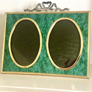 May include: A rectangular double photo frame featuring a green faux marble finish with gold-toned metal trim. The frame contains two oval-shaped openings and is topped with a decorative metal bow ornament at the center of the top edge.
