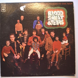 May include: A vintage vinyl record album cover featuring the band Blood, Sweat & Tears. The cover art shows a group of people, mostly men, sitting and standing in a posed group photo. The album title is in large, colorful letters and the words "A Father to the Man" are printed in smaller letters above the band name.