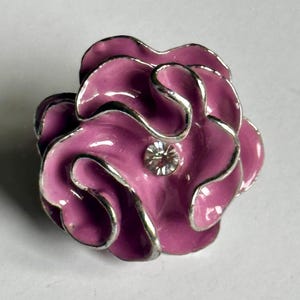 May include: A close-up of a flower-shaped brooch. The brooch features a vibrant pink enamel finish with silver-colored trim outlining each petal. A clear, sparkling rhinestone is set in the center of the flower. The brooch is likely made of metal.