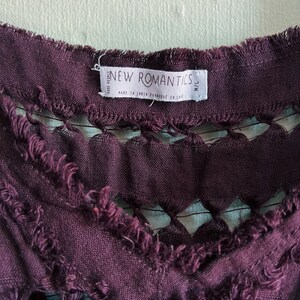 Op de afbeelding: Een close-up van een paars stoffen label met de tekst "NEW ROMANTICS" en "Made in India Fabricated en card." Het label heeft een rafelige rand.