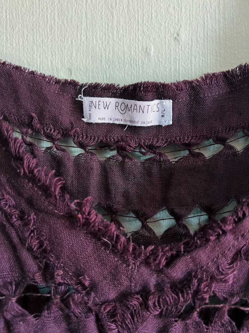 Op de afbeelding: Een close-up van een paars stoffen label met de tekst "NEW ROMANTICS" en "Made in India Fabricated en card." Het label heeft een rafelige rand.