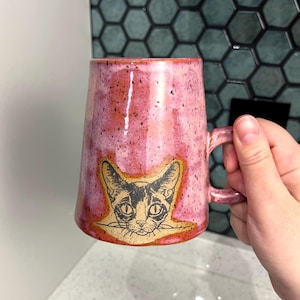 May include: A pink ceramic mug with a cat illustration. The mug has a speckled finish and a handle. The cat illustration is black and white with a tan outline. The mug is held by a hand.