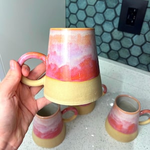 May include: Handmade ceramic mugs with a unique design. The mugs feature a vibrant gradient of pink, orange, and white on the upper portion, with a solid beige base. Each mug has a matching handle. The image shows multiple mugs.