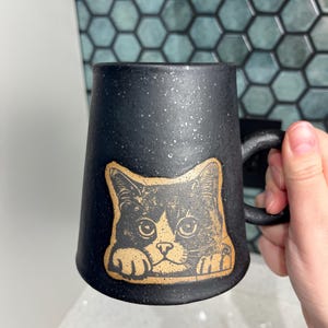 May include: Black ceramic mug with a speckled finish, featuring a cat illustration in tan and black. The cat's face and paws are visible, adding a playful touch to the mug's design. The mug has a curved handle.