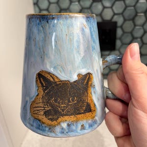 Może przedstawiać: Ceramiczny kubek z niebiesko-białą glazurą, z brązową ilustracją kota. Kubek ma uchwyt i lekko rozszerzający się kształt. Ilustracja kota jest wyśrodkowana z przodu kubka.