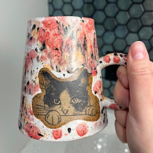 May include: A ceramic mug with a cat illustration. The mug has a white base with red and orange floral patterns. A black and white cat graphic is centered on the mug. The handle and rim have red and black accents.