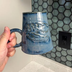 Könnte beinhalten: Ein Keramikbecher mit glänzender blauer Glasur und einem eingeprägten T-Rex-Schädel-Design auf der Vorderseite. Der Becher hat einen abgerundeten Henkel und eine leicht konische zylindrische Form mit glatter Oberfläche und dunklen blauen Akzenten.