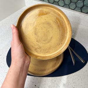May include: Two handmade ceramic plates with a speckled, golden-yellow glaze. The plates have a slightly raised rim and a rustic, artisanal appearance. The plates are stacked on top of each other.