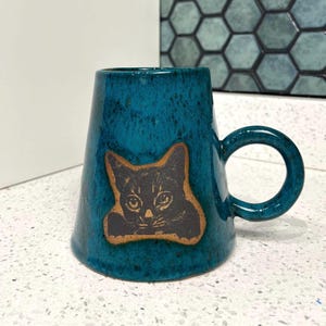 Może przedstawiać: Turkusowy ceramiczny kubek z ilustracją czarnego kota. Kubek ma unikalny kształt, zaokrąglony uchwyt i teksturowane wykończenie. Ilustracja kota jest obramowana brązową obwódką.