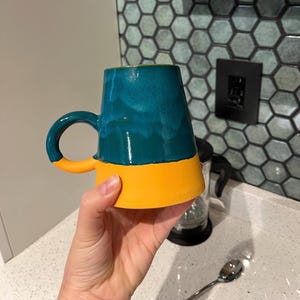 May include: A teal and yellow ceramic mug with a rounded handle. The mug's upper portion is teal with a glossy finish, while the bottom half and handle are bright yellow. The mug is held in a hand, showcasing its unique color combination and design.