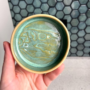 Puede incluir: Un pequeño plato redondo de cerámica con interior turquesa y borde marrón claro. El plato presenta un diseño en relieve detallado de un cráneo de dinosaurio. El plato se sostiene en una mano, mostrando su tamaño y artesanía.