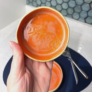 May include: Orange ceramic dish with a raised triceratops skeleton design. The dish has a rounded shape with a light yellow rim. A second dish is visible in the background. The dish is held by a person's hand.