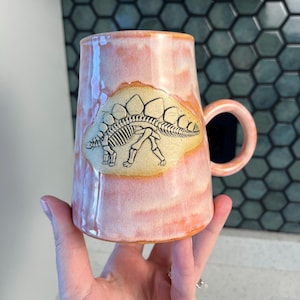 May include: A tall, pink ceramic mug with a handle. The mug features a Stegosaurus skeleton design on a tan patch. The mug has a unique shape and a glossy finish, perfect for coffee or tea.