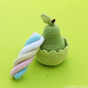 Puede incluir: Un juguete de crochet con forma de pera en color verde y blanco. La pera está sentada sobre una superficie verde junto a un juguete de cuerda trenzada de crochet multicolor.