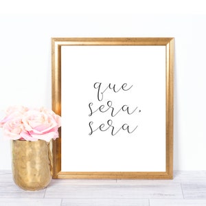 Könnte beinhalten: Ein goldfarbener gerahmter Druck mit dem Text "que sera, sera" in schwarzer Kursivschrift. Der Druck liegt auf einer weißen Holzoberfläche mit einem Strauß rosa Rosen in einer goldenen Vase.