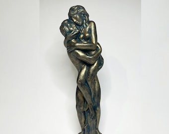 Puede incluir: Escultura de color bronce que representa dos figuras abrazándose. Las figuras están entrelazadas, una de ellas sosteniendo a la otra. La escultura tiene una base detallada y está sobre un fondo blanco. La estatua mide aproximadamente 30 cm de altura.