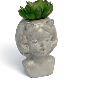 May include: A gray concrete planter shaped like a girl's bust with a green succulent plant growing out of the top.