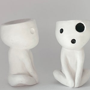 Puede incluir: Dos macetas de cerámica blanca con forma de pequeñas figuras humanas. Una maceta tiene ojos y boca pintados de negro. La otra maceta es lisa.